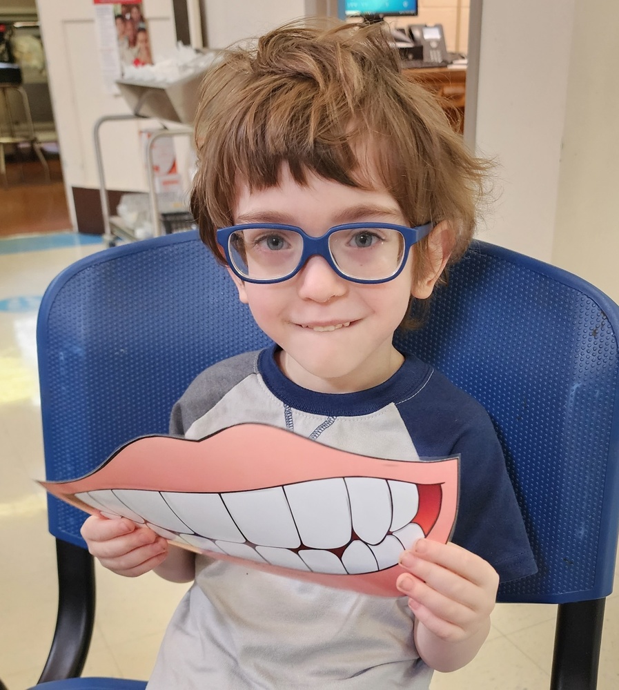 A preschool student with blue glasses smiles while holding up a large cutout of a smiling mouth