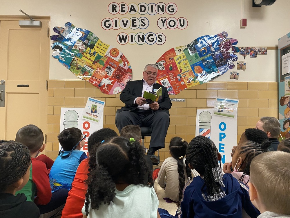 Carter Bank CEO sits in a barber chair reading a book to students who are seated around him on the floor. On the wall behind him are two wings made out of book covers with the phrase "reading gives you wings" above them.