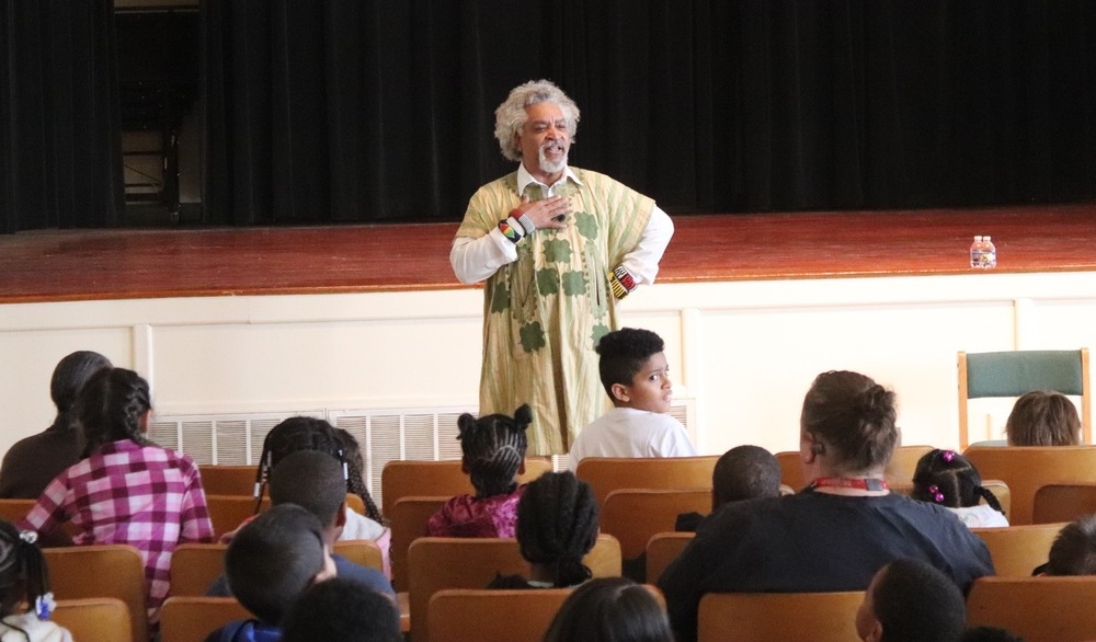 Storyteller Fred Mott stands with his hand on his chest in front of seated elementary students