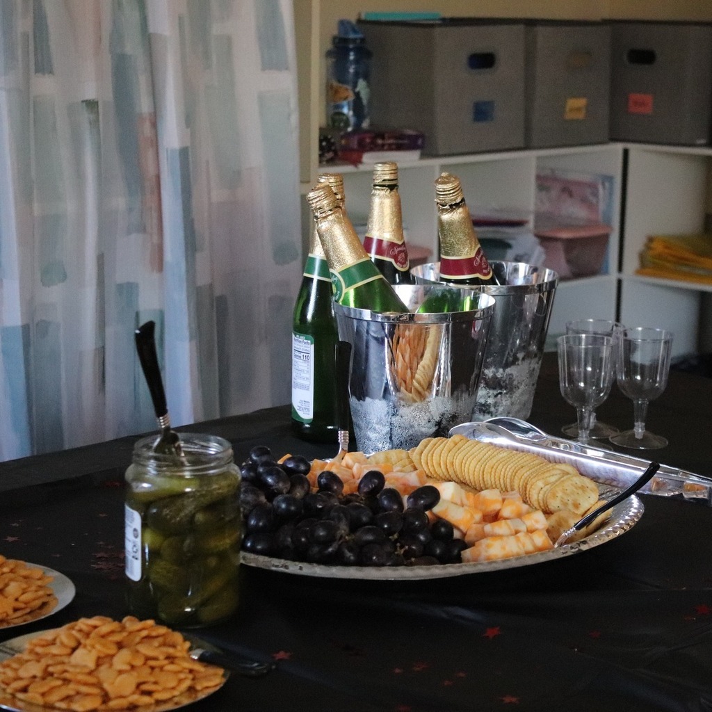 A table covered in a black tablecloth holds silver buckets with bottles of sparking grape juice, a tray of cheese, crackers, and grapes, and other snacks