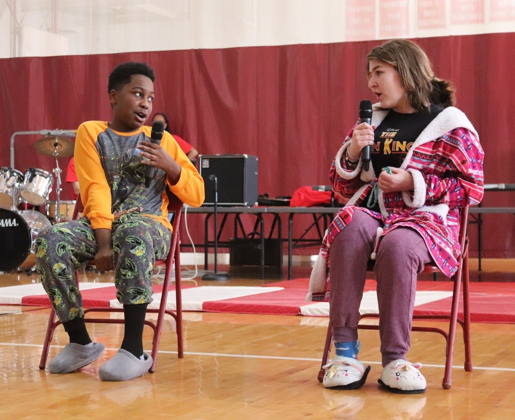 two students dressed in slippers, pajamas, and bathrobes sing into microphones while sitting in chairs during the MMS talent show