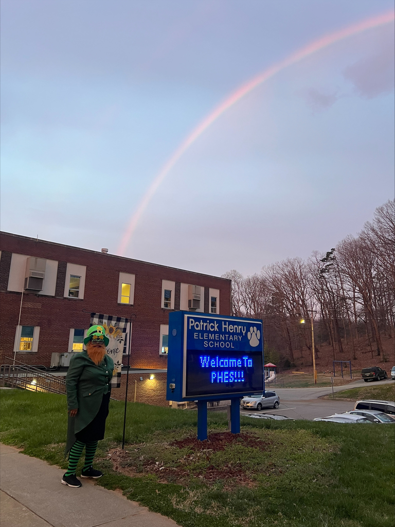 A rainbow arches over the sky and ends at Patrick Henry Elementary School, where a teacher stands in a leprechaun costume on the front sidewalk