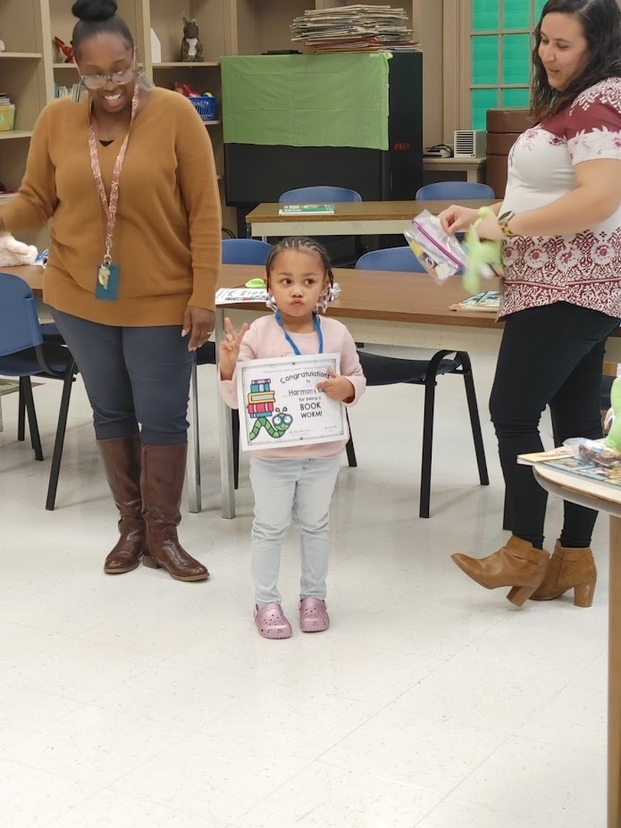A preschool girl holds up the peace sign with one hand and holds a reading certificate in the other. Two teachers on either side of her smile. 