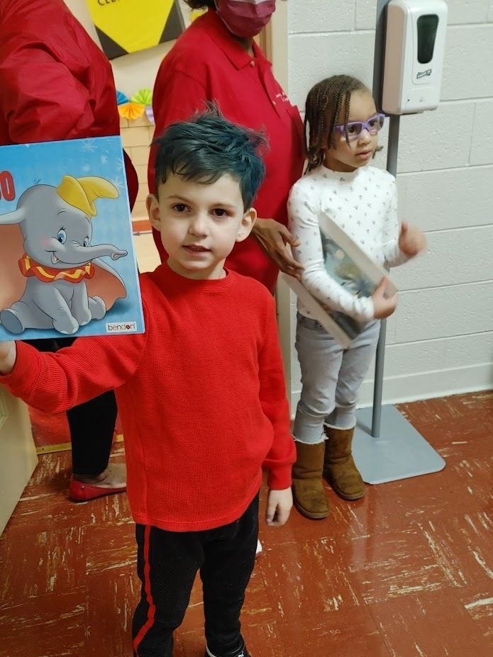 A preschool student with blue hair and a red shirt holds up a book with Dumbo on the back cover