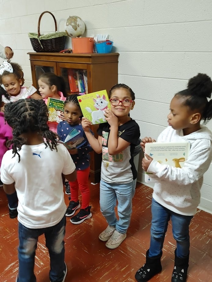 Preschool students stand with their books. One holds a book up to their face and smiles at the camera.