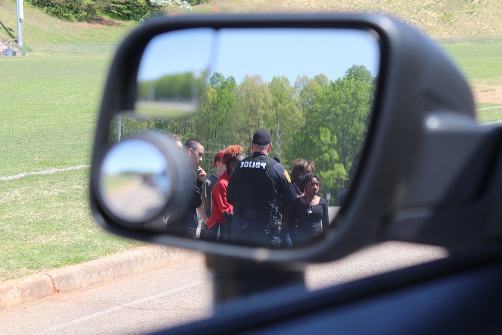 A police officer and high school students are shown in the reflection of a car's side view mirror